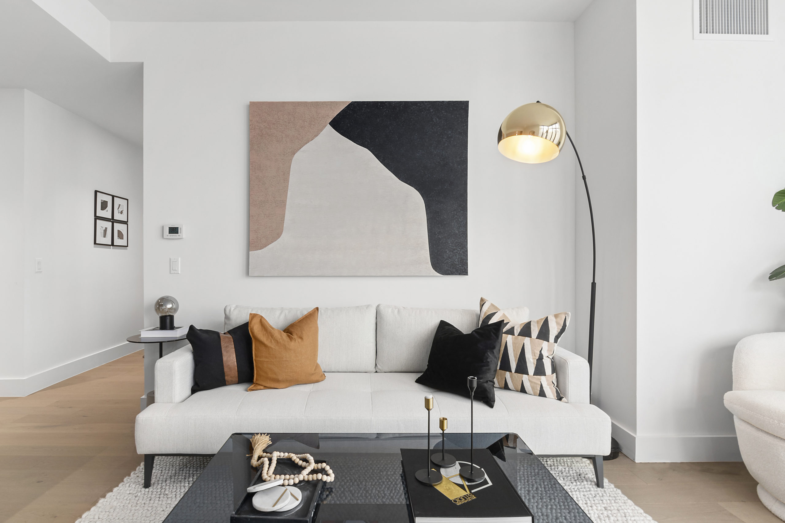 Modern living room with white couch with black and brown pillows, large contemporary painting above on white walls, dark glass coffee table on beige area rug on wood floors and contemporary floor lamp