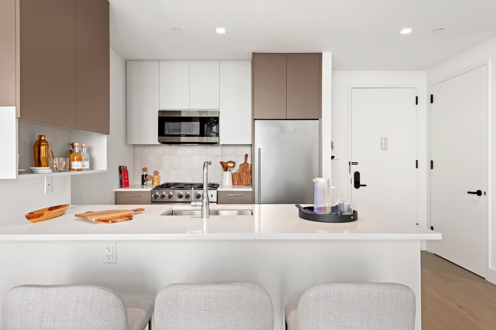 Modern kitchen near entrance with light grey and tan cabinetry, white counters with seating, stainless steel applicances and wood floors