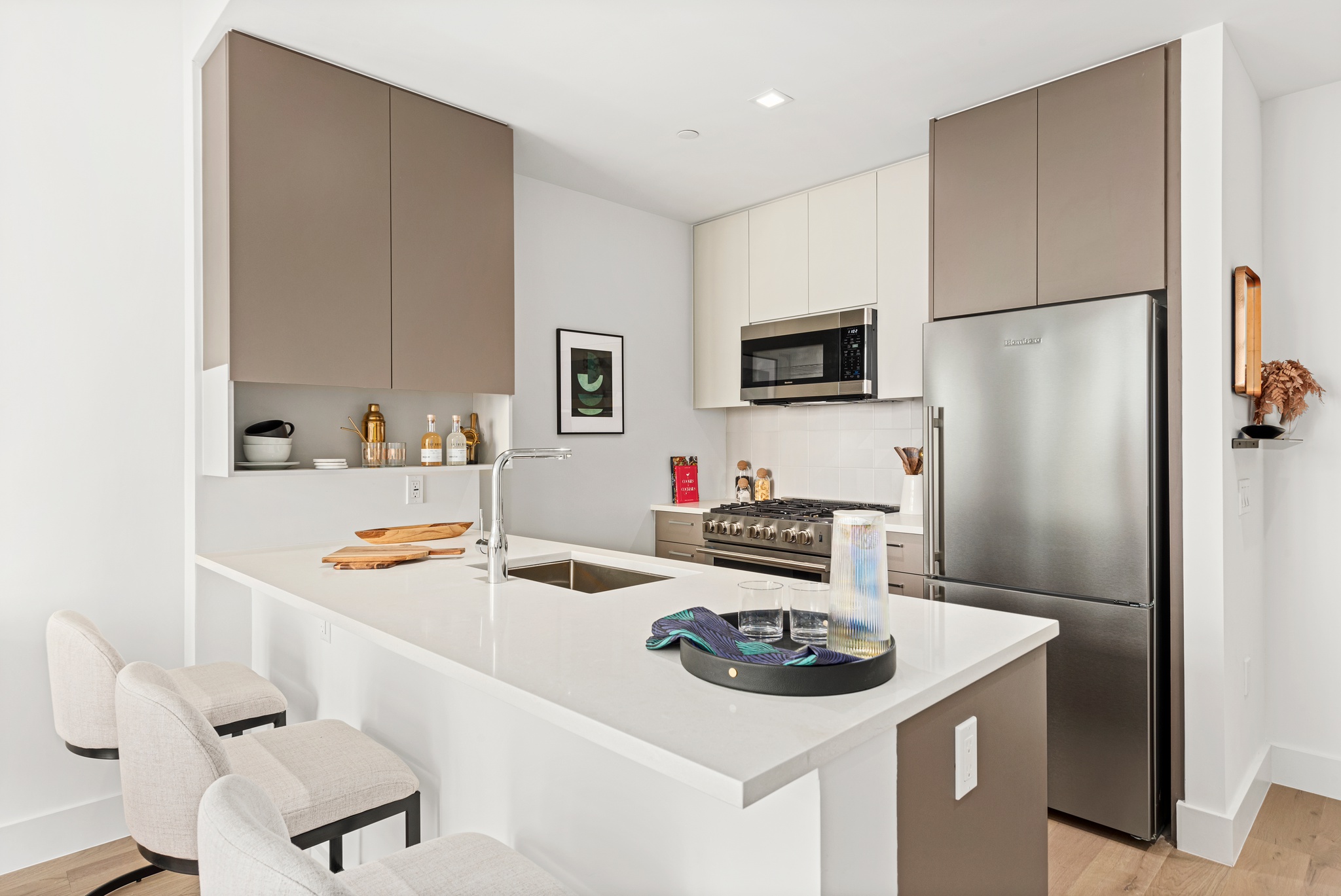 Modern kitchen with light grey and tan cabinetry, white counters, stainless steel applicances, wood floor and counter seating with white and black bar stools