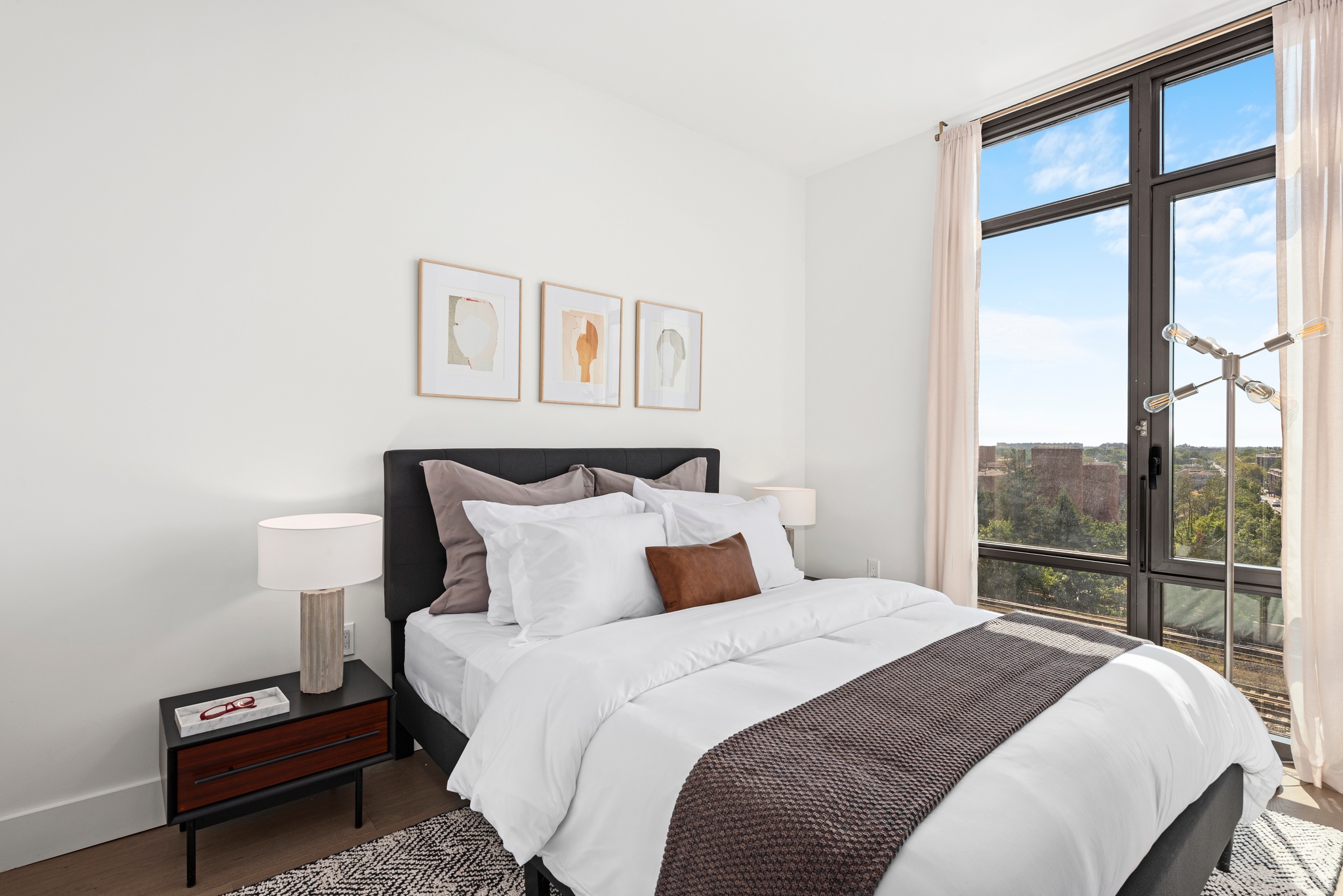 Bright bedroom with white walls, white and brown bedding, dark headboard with tryptic art above, large windows and matching drawer night stands with lamps