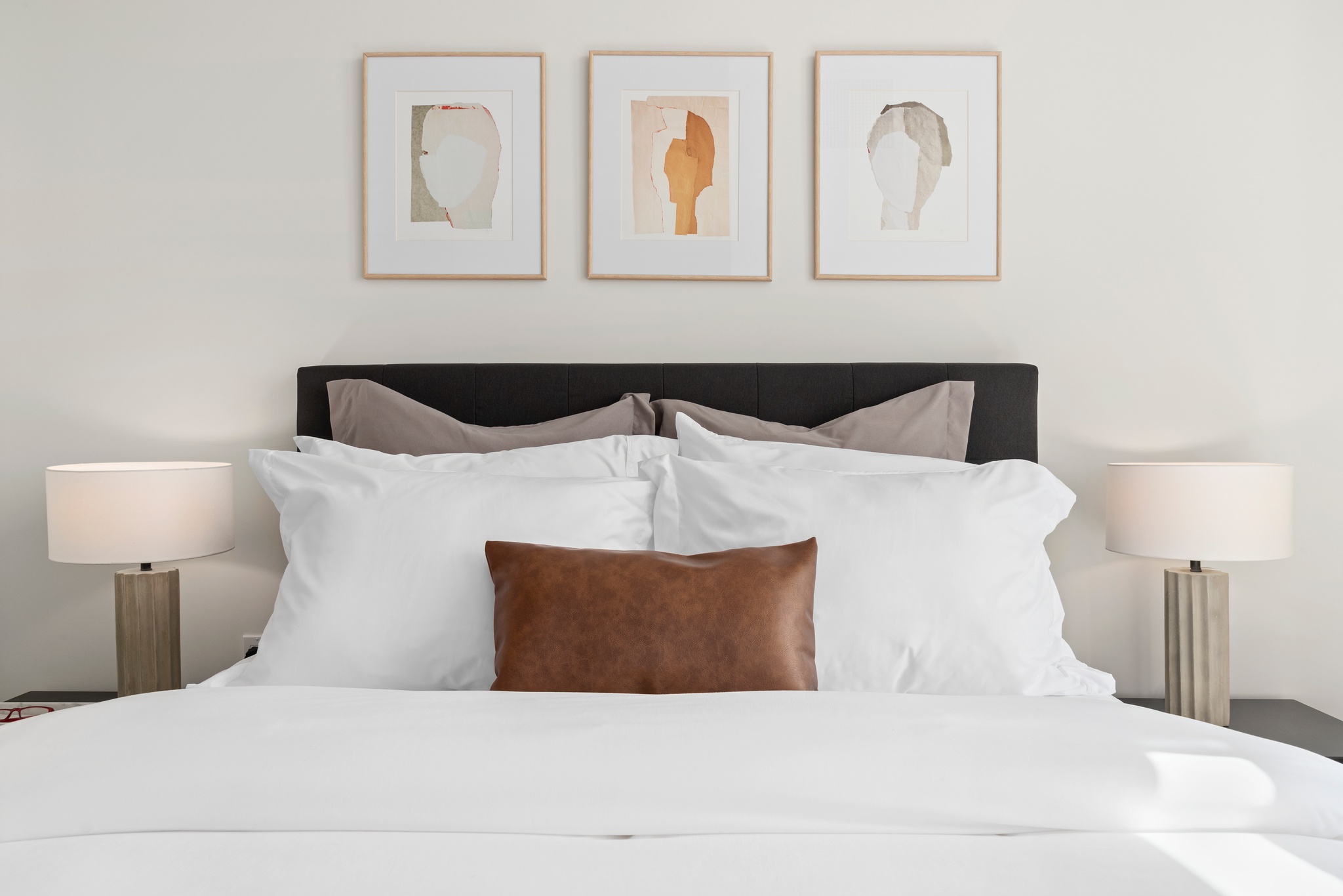 Bright bedroom detail showing white walls, white and brown bedding, dark headboard with tryptic art above, dark headboard and lamps on matching nightstands