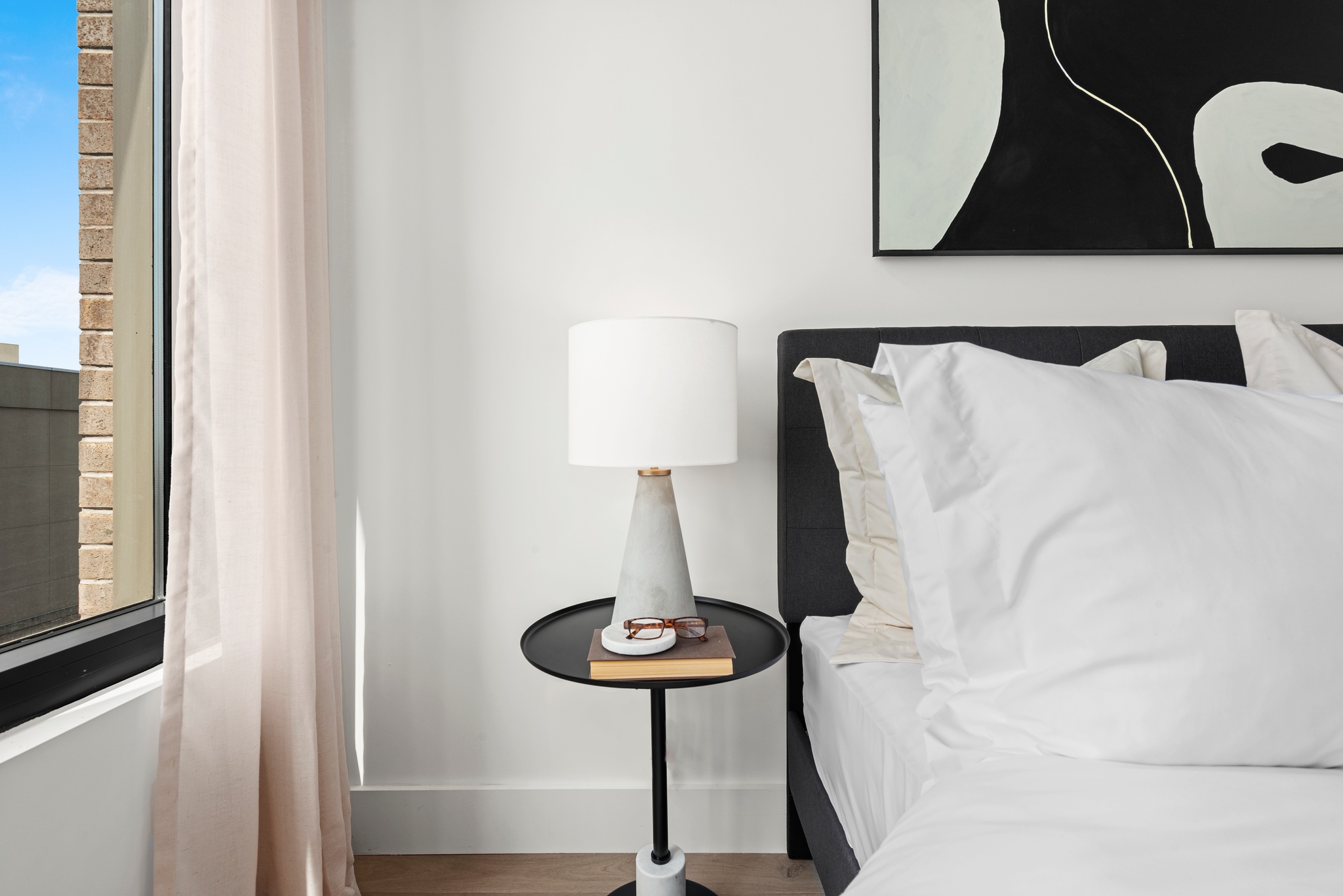 Bedroom detail of corner of dark bed frame with white bedding, side table with lamp, book and glasses and partial window with light curtains