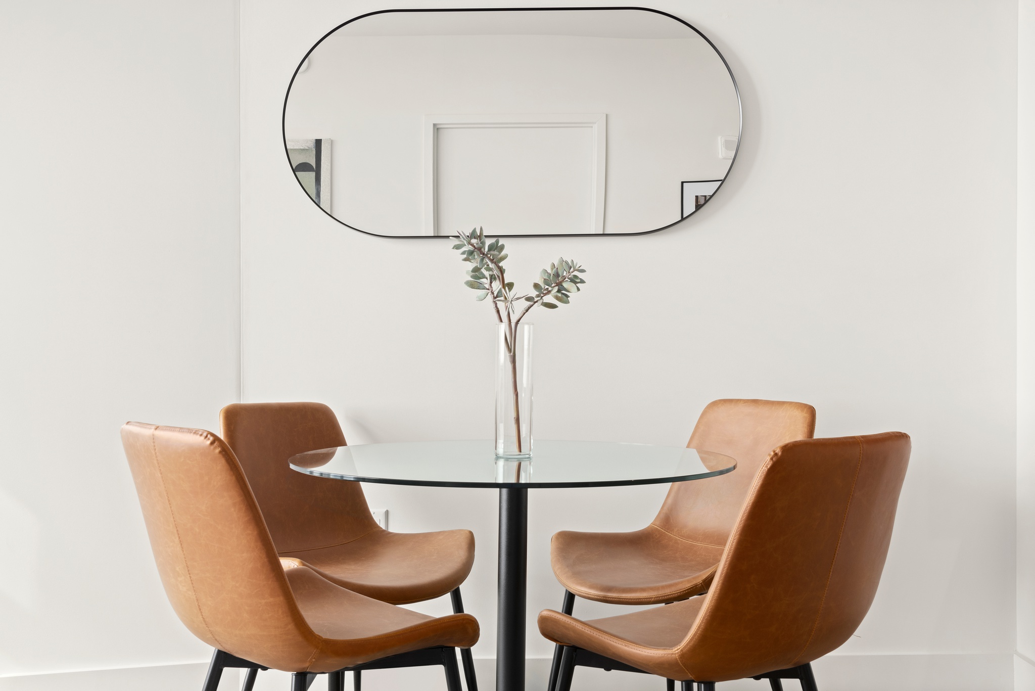 Living room detail of round glass eat in table with camel leather chairs and oval mirror above on white wall
